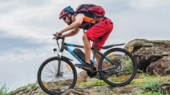Person riding a VARUN M27-1 Electric mountain bike on rocky terrain with a backpack and helmet.