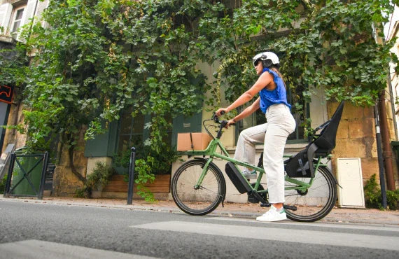 Woman riding a green electric cargo bike with rear seat on a city street side view