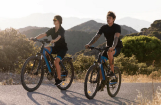 Two people riding blue electric bikes outdoors on a scenic mountain road, Pogo Cycles
