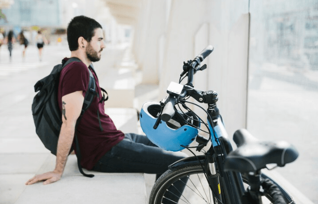 Electric bike parked cityside, blue helmet on handlebars, man with backpack resting nearby